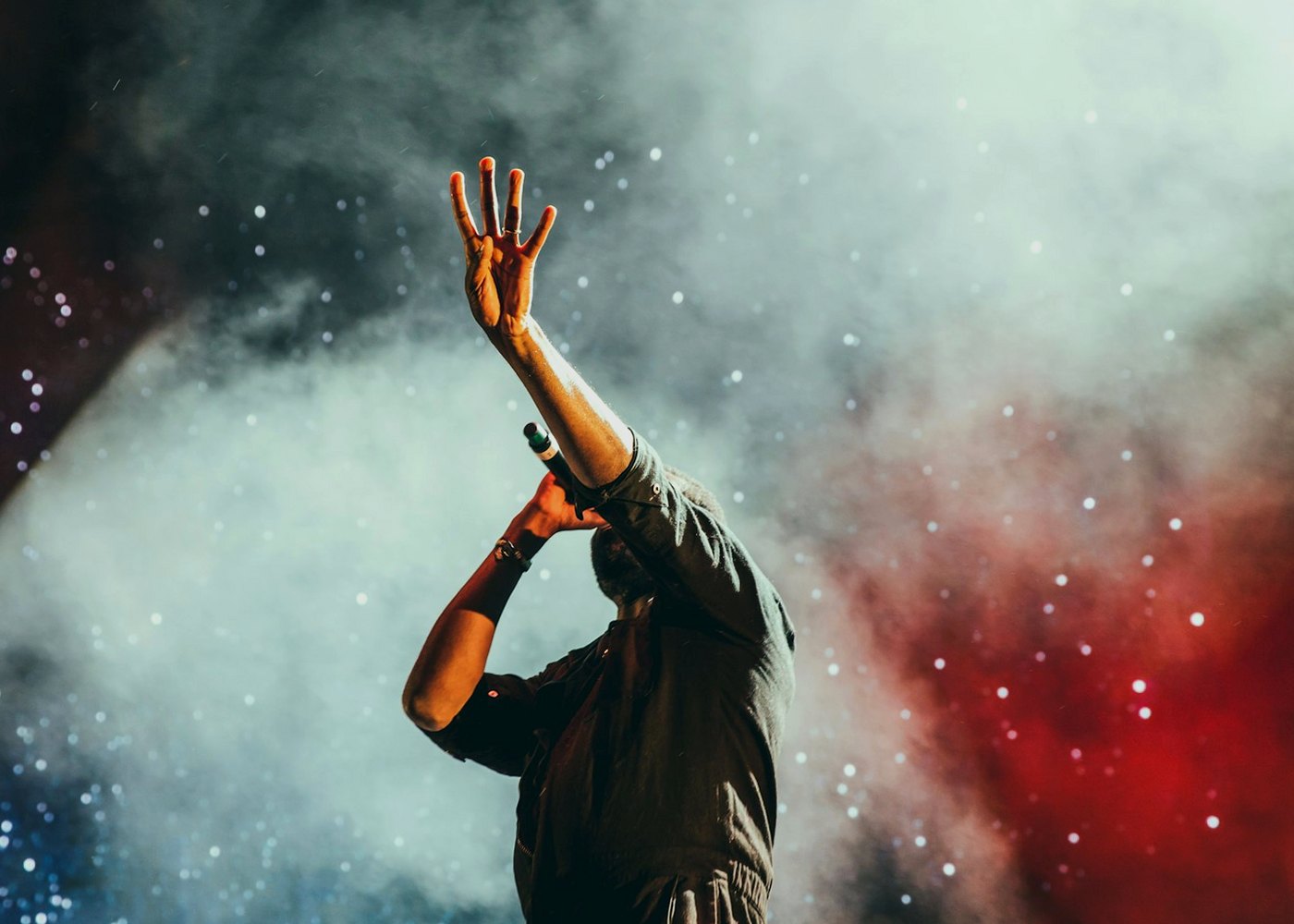 Performer on stage holding a microphone with one hand raised in the lights